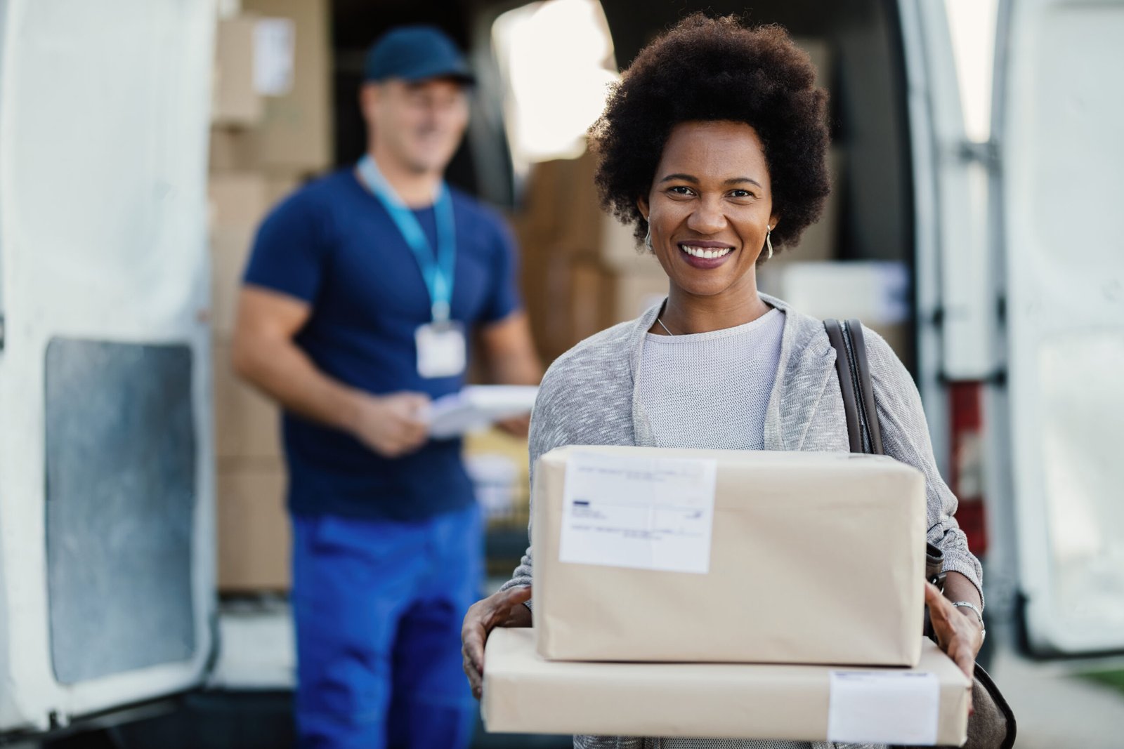 Portrait of happy black woman getting her packages delivered and looking at camera. A courier is standing in the background.